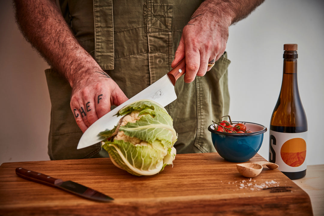 Man using a chef’s knife to chop fresh cauliflower on a wooden cutting board.