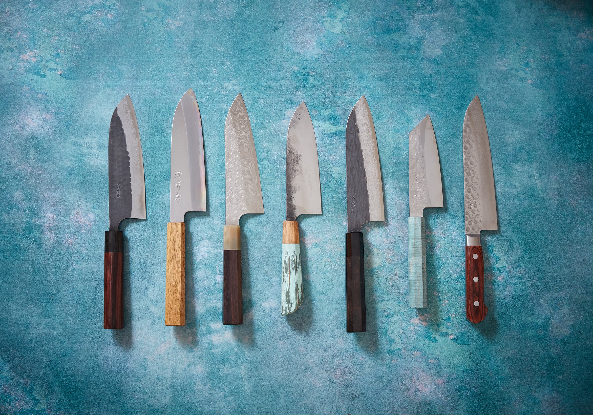Seven handmade Japanese santoku kitchen knives on a blue background