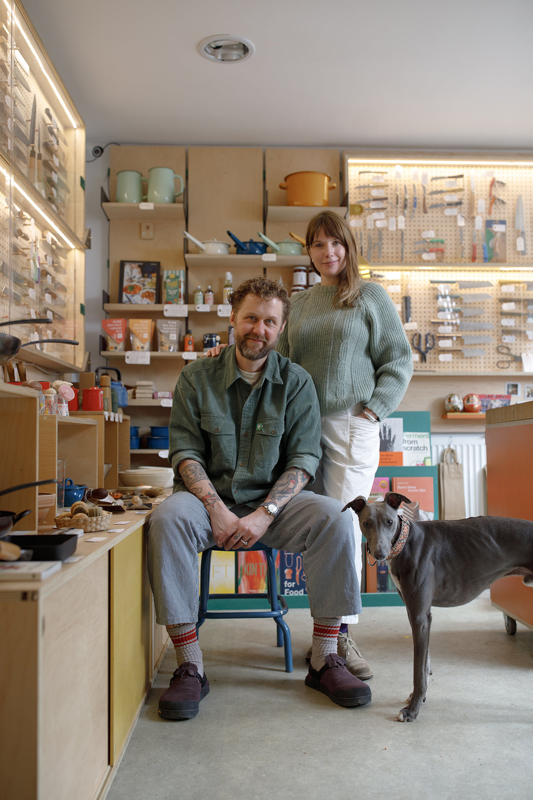 Man sitting on a chair with a woman standing behind him and a dog in a kitchenware store setting. Japanese knives in a cabinet behind them.