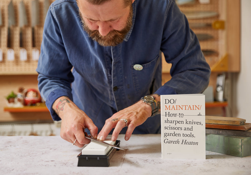 Man sharpening a knife with a book titled 'Do Maintain!' on a table.